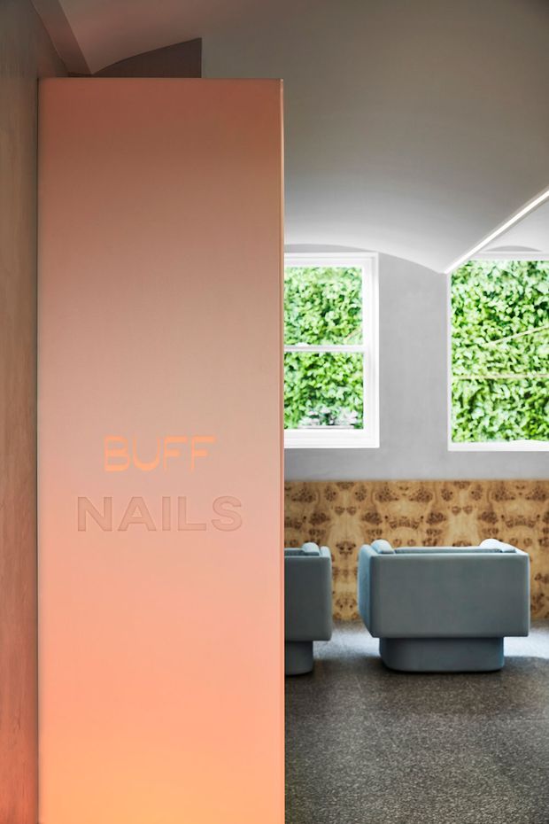 Interior view of a contemporary nail salon, looking past a tall peach-pink partition or entry panel embossed with “BUFF NAILS.” The salon contains muted blue-grey upholstered chairs, a dark carpeted floor, a textured brown timber feature wall, white-framed windows, and a curved vaulted ceiling with linear lighting. Green foliage is visible outside the windows.