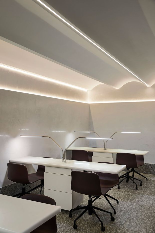 A refined contemporary commercial interior featuring several white worktables with integrated task lighting, dark upholstered swivel chairs, and central white storage pedestals. The room has softly curved ceiling details with concealed LED strip lighting, pale textured wall surfaces, and a speckled terrazzo-like floor.