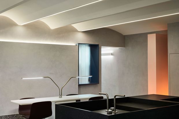 A sophisticated contemporary kitchen with a large dark island and integrated sink, pale stone or plaster-finished walls, a white peninsula or work surface, dark bar stools, and sculptural brushed-metal tapware. The ceiling features recessed linear lighting and curved illuminated ceiling transitions, while a warm orange-lit vertical niche adds contrast.