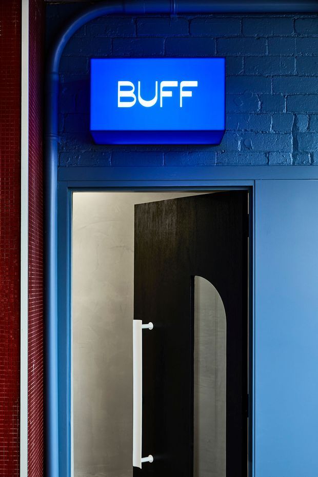 Close-up of a bold commercial entrance featuring a vivid blue painted brick wall, exposed blue pipework, a glowing blue 'BUFF' sign, and a black door with a tall arched inset and white pull handle. A red textured wall edge contrasts with the blue industrial-style fit-out.