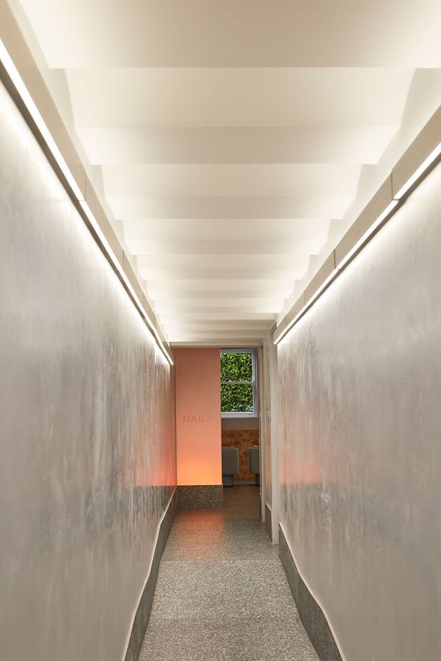 A narrow, long interior corridor with softly textured pale walls, continuous linear wall lighting, a stepped white ceiling with recessed beams, and a grey speckled floor. A warm peach-toned doorway or partition at the far end leads toward a small room with a window and greenery visible outside. The corridor has a clean, contemporary, softly illuminated atmosphere.