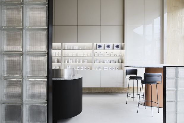Bright, minimalist retail or showroom interior with a monochromatic white built-in display wall, neatly arranged products, a black cylindrical counter, and a small bar-height seating area. Translucent glass-block partitions frame the space, while pale flooring and glossy cabinetry create a clean contemporary atmosphere.