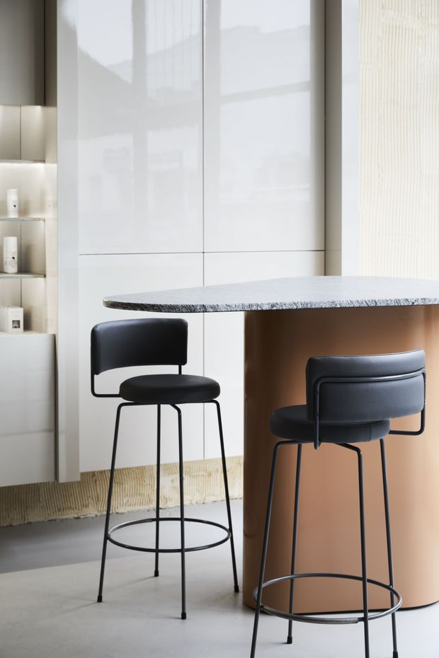 A refined modern kitchen or hospitality-style breakfast counter with a pale stone or engineered-stone benchtop supported by a rounded warm brown base. Two black upholstered bar stools sit on a smooth grey concrete-like floor. The background features tall glossy white cabinetry, softly illuminated open shelving, and vertical textured wall panelling, creating a clean, minimalist interior.