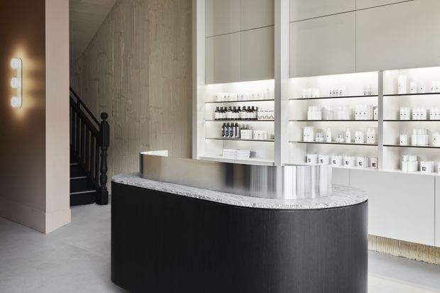 A refined commercial retail or reception space featuring a large curved dark timber-toned counter with a pale stone benchtop, illuminated white built-in shelving displaying small products, and a sculptural staircase with dark balustrade in the background. The space has a restrained, monochromatic palette and integrated architectural lighting.
