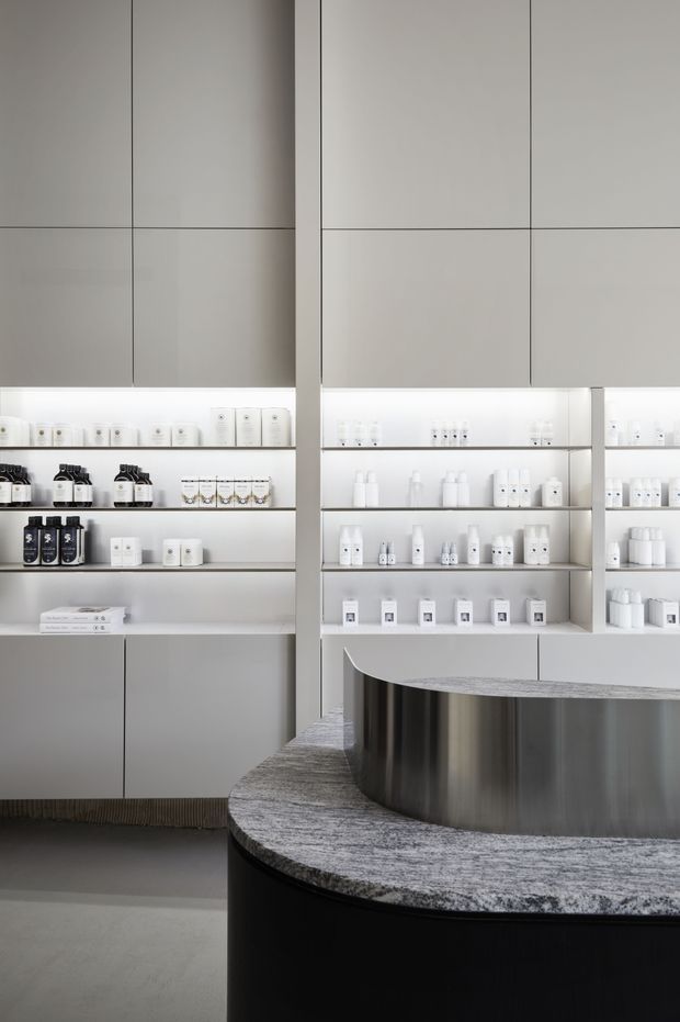 A minimalist retail or commercial interior with a sculptural curved reception or display counter in dark cabinetry, a grey stone-like benchtop, and integrated brushed metal detailing. The surrounding walls feature pale grey full-height cabinetry and open illuminated shelving displaying small products. The space is monochromatic, clean-lined, and softly lit.