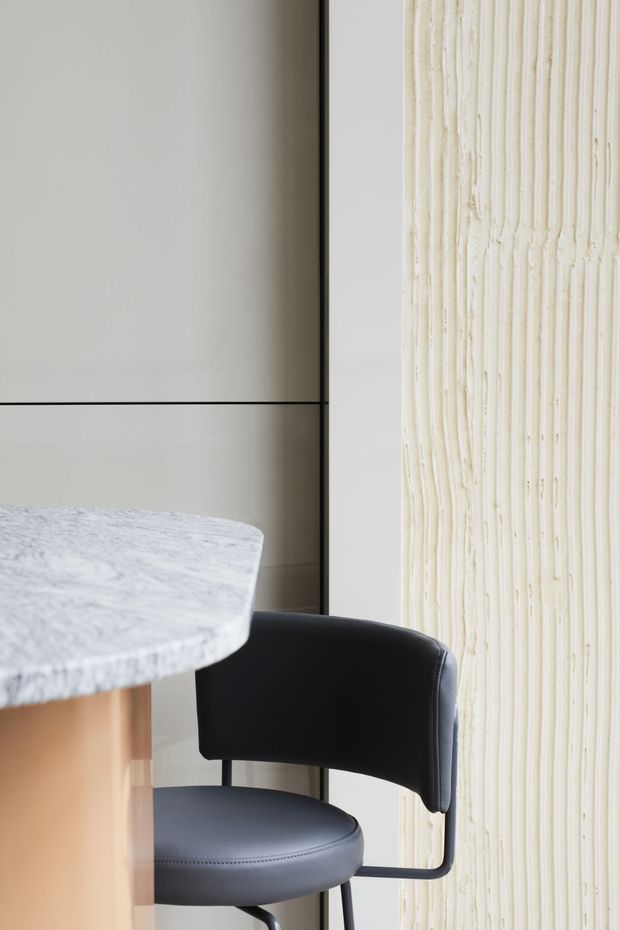 Close-up interior detail of a refined dining or kitchen seating area, showing a black upholstered chair beside a rounded pale marble benchtop or table. The background combines large light neutral panels with a vertically textured cream feature surface, creating a minimalist, contemporary composition.