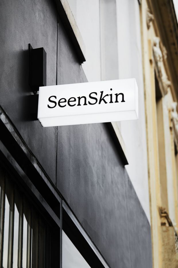 Close-up exterior view of a contemporary commercial storefront sign reading “SeenSkin,” mounted perpendicular to a dark charcoal facade. The sign is a simple illuminated white rectangular lightbox, with black-framed glazing below and a contrasting older pale facade visible to the side.