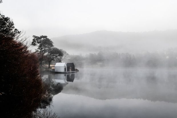 A quiet, mist-covered lake with a small minimalist cabin or boathouse positioned on the far shoreline and reflected in the still water. Dense wooded hills and trees fade into the fog, creating a monochromatic, atmospheric landscape scene.