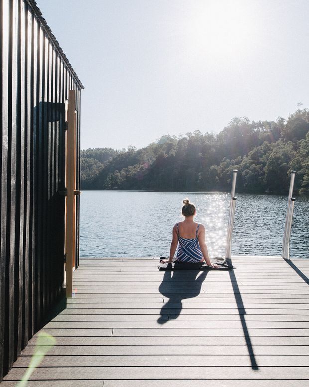 A sunlit timber waterfront deck beside a calm lake, viewed from an open dark-clad structure. A person sits at the edge facing the water, with a metal swim ladder and forested shoreline beyond. The image has a serene, outdoors-oriented architectural photography style with strong shadows and reflective water.
