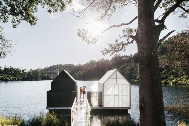 A sunlit lakeside scene showing two small geometric structures positioned over the water at the end of a narrow timber footbridge: a dark-clad cabin-like volume and a translucent glazed or polycarbonate gabled pavilion. Dense trees surround the lake, with people standing on the bridge. The composition highlights a contemporary waterside architectural installation and its natural setting.