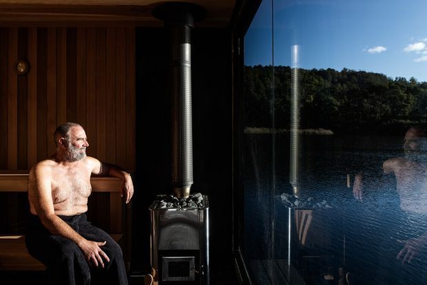 A compact timber-lined sauna beside a large glazed wall overlooking a calm lake and forested shoreline. A shirtless person sits on a wooden bench next to a black wood-burning sauna stove with a vertical flue. The scene combines dark natural timber, dramatic sunlight, reflective glass, and an expansive waterside view.
