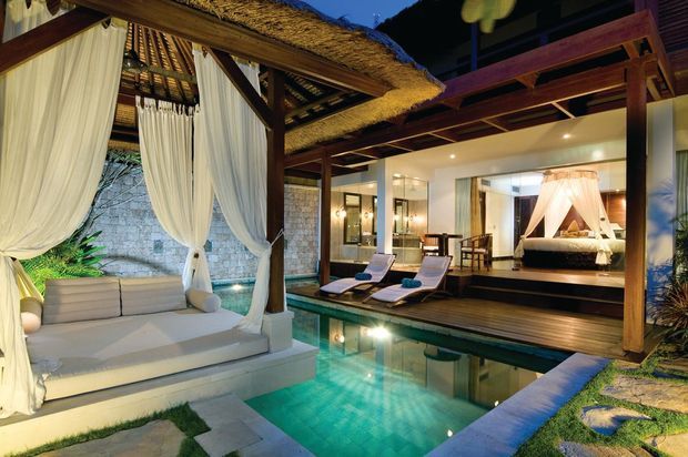 Luxury tropical resort-style outdoor living area with a private turquoise plunge pool, a timber-framed covered pavilion, a thatched roof, flowing white drapery, built-in daybed, and loungers. The pavilion opens toward an illuminated bedroom with a prominent four-poster bed and canopied curtains. Natural stone walls, timber decking, lush planting, and warm ambient lighting create an intimate, tranquil setting.