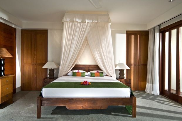 A spacious, warmly lit bedroom centered on a four-poster-style bed with flowing white canopy curtains. The room features dark timber built-in wardrobes and cabinetry, matching bedside tables and lamps, white walls, large timber-framed glazed doors or windows with sheer curtains, and a grey carpeted floor. Colourful cushions and floral accents add small decorative highlights.