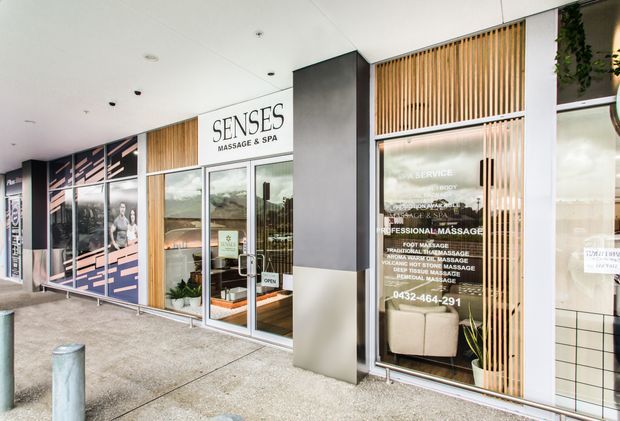 Street-level exterior view of a contemporary commercial massage and spa storefront. The facade features full-height glazing, aluminium-framed glass doors and windows, dark grey wall panels, vertical timber-look screening, and prominent business signage.