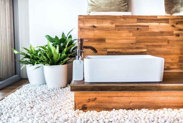 Close-up of a contemporary bathroom vanity featuring a white rectangular vessel basin, chrome tapware, and warm timber wall and vanity joinery. White decorative pebble flooring, potted green plants, and a large window contribute to a bright, natural aesthetic.