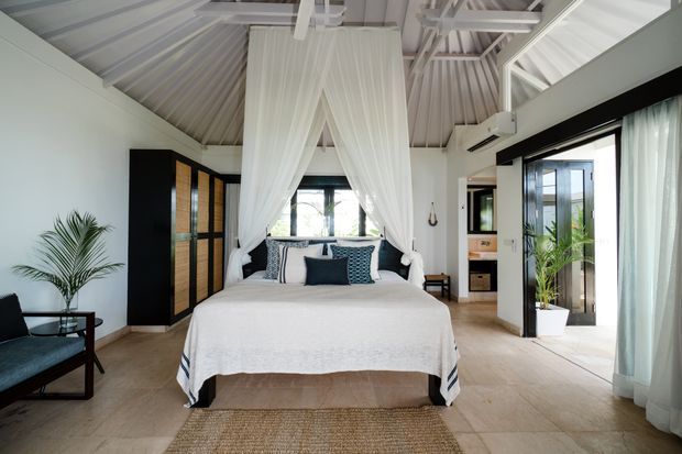 Bright, airy bedroom with a central four-poster bed draped in sheer white fabric, exposed white vaulted roof structure, pale tiled flooring, black-framed glazing, dark timber joinery, and tropical planting. The room opens toward a compact adjoining bathroom or dressing area and has a relaxed resort-like character.