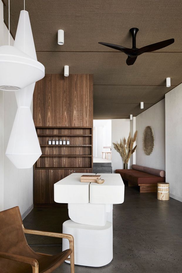 A refined open-plan living and dining space with a sculptural white dining table and pendant lights, dark timber built-in shelving, a brown low-profile sofa, leather armchair, and concrete flooring. The room features a dark textured ceiling, minimalist cylindrical wall lights, a ceiling fan, neutral plaster walls, and warm natural styling with dried grasses and wall decor.