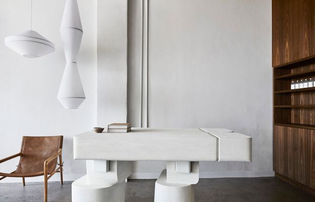 A refined minimalist interior featuring a sculptural white stone or concrete desk with chunky pedestal supports, matching pendant lights, a brown leather sling chair, and a dark timber built-in shelving wall. The room has white textured walls and a dark polished floor, creating a quiet, gallery-like workspace.