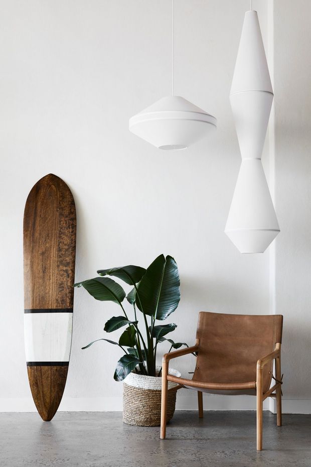A minimalist interior vignette with white walls and a grey concrete floor. Two sculptural white pendant lights hang above a brown leather and timber armchair, beside a tall broad-leaf potted plant in a woven basket. A vertically displayed wooden surfboard with a white painted stripe leans against the wall.
