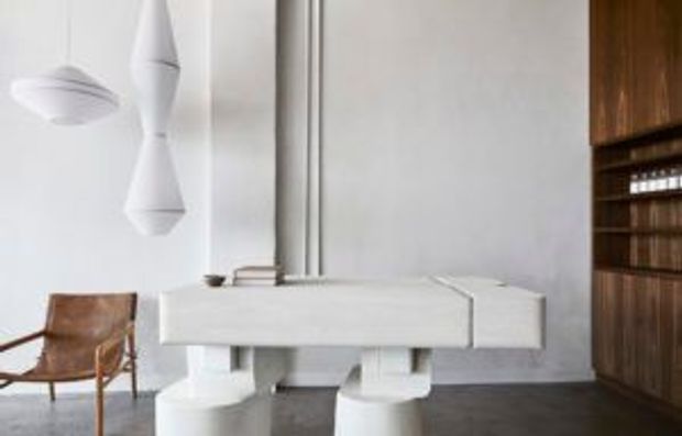 Minimalist interior featuring a white sculptural desk or console with substantial pedestal supports, a brown leather lounge chair, sculptural white pendant lights, and a dark timber built-in shelving wall. The palette is predominantly white with warm wood and leather accents.