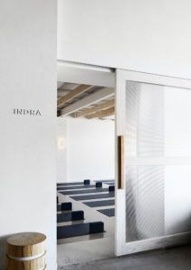 A minimalist white interior viewed through a wide sliding doorway, with a partially visible room beyond featuring exposed timber ceiling beams and bold black-and-white geometric floor or seating elements. The foreground has a small round wooden stool, while the doorway includes a warm timber handle and white-framed panels.