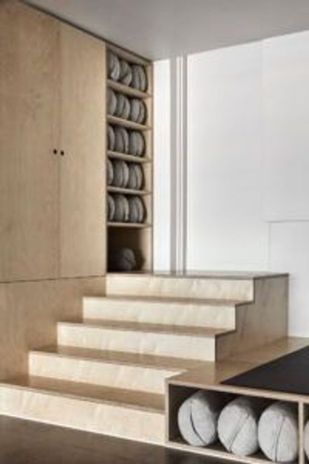 A minimalist interior stairway with pale timber built-in cabinetry and shelving integrated around the staircase. The open shelves display stacked round objects, while white walls, light stair treads, and clean linear detailing create a restrained, contemporary look.