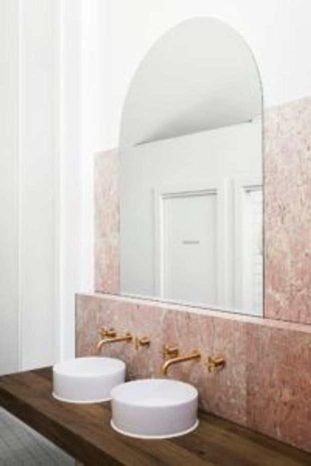Bright contemporary bathroom vanity with two round white vessel basins set on a dark timber benchtop, paired with wall-mounted brass tapware. A pink-toned stone or terrazzo backsplash extends behind the vanity, with a large arched mirror and white paneled wall details.
