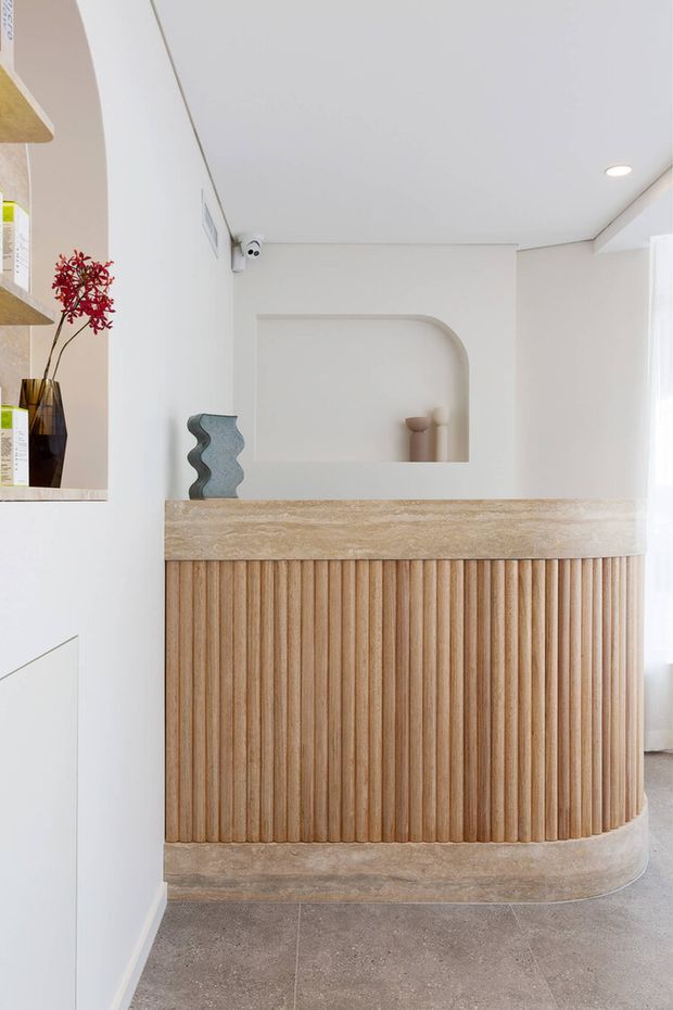 Bright, refined reception or entry area featuring a curved built-in timber slatted counter with a beige stone or travertine benchtop and matching curved base. White walls include a softly arched recessed display niche, with subtle decorative objects and integrated shelving. Large-format grey flooring and sheer window light create a calm, contemporary atmosphere.
