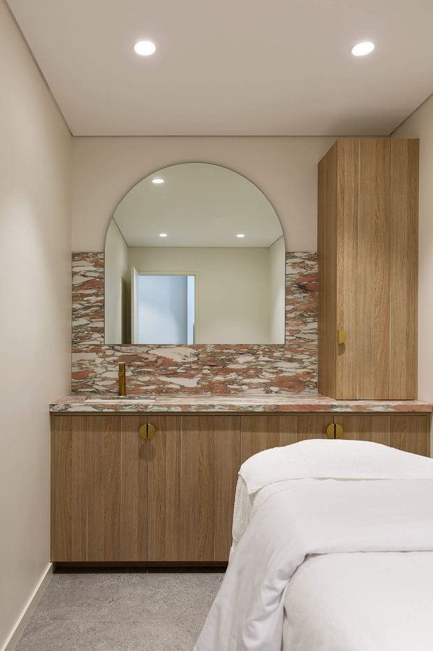 A refined bedroom vanity area featuring a timber-grain built-in cabinet with integrated basin and brass tapware, a dramatic patterned stone splashback and benchtop, and a large arched mirror. A bed is visible in the foreground, confirming the room as a bedroom. The space has warm neutral walls, recessed ceiling lighting, and a soft grey carpet.