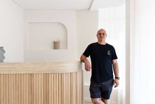 Bright minimalist interior featuring a curved reception or service counter with a textured pale stone benchtop and vertical light timber slat joinery. A man stands beside the counter, with an arched wall niche and soft sheer window treatments in the background.
