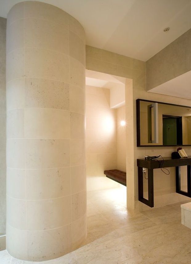 Warm, refined bathroom interior featuring pale stone or limestone wall and floor finishes, a curved tiled feature wall, recessed shower or bathing area with a dark timber bench, and a dark-framed wall mirror above a slim black vanity console. Soft recessed lighting creates a calm, luxurious atmosphere.
