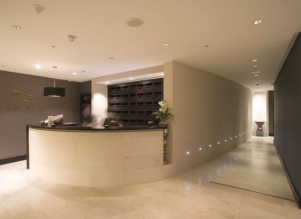 A refined reception lobby with a curved pale stone concierge desk, dark feature wall, pendant lighting, recessed ceiling lights, and a long illuminated corridor leading deeper into the building. Built-in mail or storage compartments and subtle decorative planting contribute to a polished commercial interior.