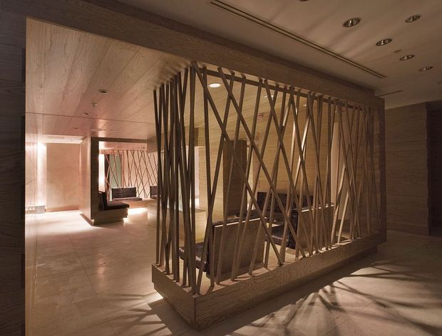 A warm, dimly lit contemporary interior with a dramatic sculptural timber screen dividing circulation and lounge areas. The screen consists of irregular diagonal timber members set within a substantial timber frame, with pale stone or tile flooring, timber wall and ceiling lining, recessed downlights, and a seating area visible beyond.