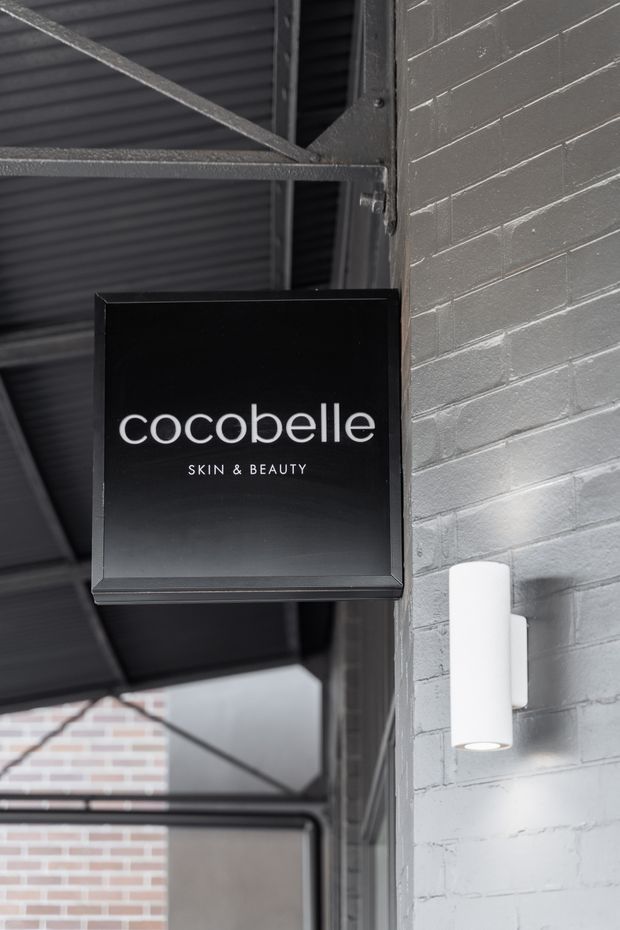 Close-up of a black rectangular sign reading “cocobelle SKIN & BEAUTY” mounted on a grey-painted brick wall beneath a dark corrugated metal canopy. A white cylindrical wall light is mounted beside it, with exposed metal framing and brickwork visible in the background, creating an industrial commercial entry or facade detail.