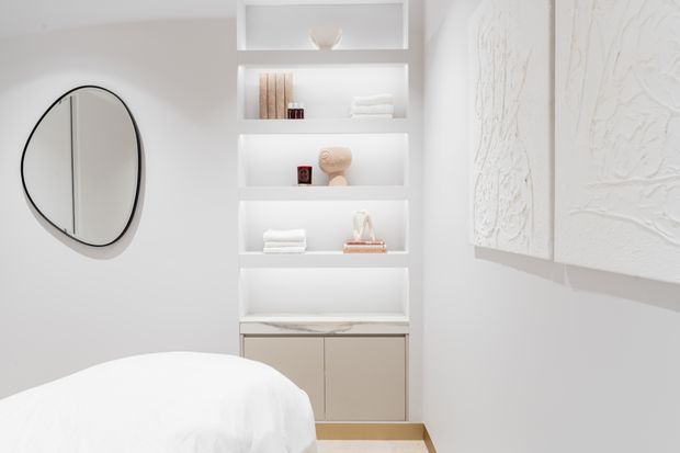 Bright, serene bedroom corner featuring a curved upholstered bed in the foreground, a round black-framed wall mirror, and a built-in white shelving unit with minimalist decor and closed lower cabinetry. A textured white wall artwork adds subtle visual interest.