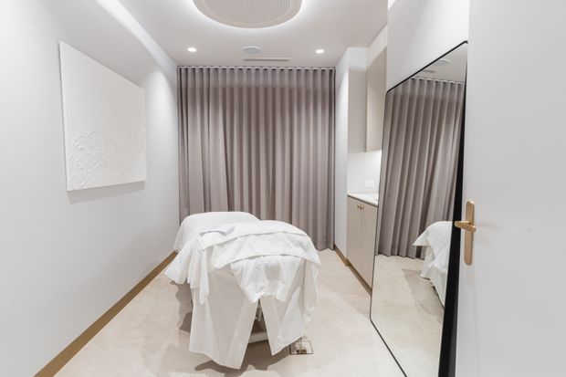 A compact, bright treatment room with a massage table draped in white linens, floor-to-ceiling taupe curtains, a pale tiled floor, white walls, recessed ceiling lights, built-in cabinetry, and a large full-height mirror on the door. The space has a clean, minimalist commercial wellness aesthetic.