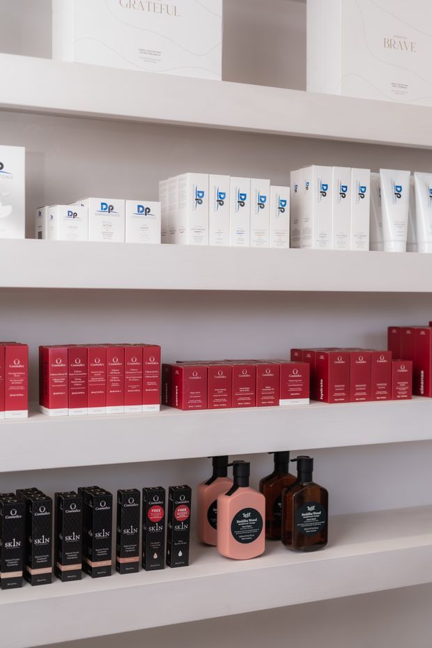 A close-up of white built-in retail display shelving stocked with neatly arranged skincare and beauty product boxes and pump bottles in white, red, black, pink, and brown packaging. The shelves are set against a soft warm-grey wall, with framed motivational artwork visible above.
