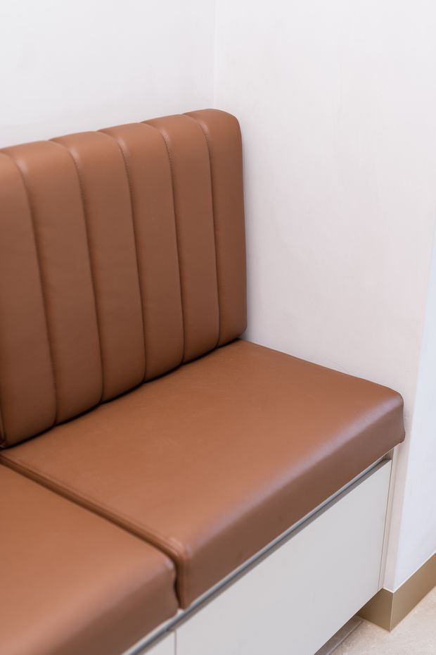 Close-up of a built-in upholstered bench seat in warm brown leather or faux leather, featuring vertically channelled padding against white walls. The seat incorporates white cabinetry below with a slim metallic trim and sits on a light floor with a beige skirting board.