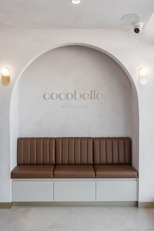 A minimalist beauty and skincare reception area featuring a built-in brown channel-tufted bench beneath a large rounded arch niche. The niche and surrounding walls are finished in soft off-white plaster, with subtle wall sconces, recessed ceiling lighting, and a visible security camera. The bench incorporates white built-in cabinetry below, while large pale stone-look floor tiles complete the calm, refined interior.