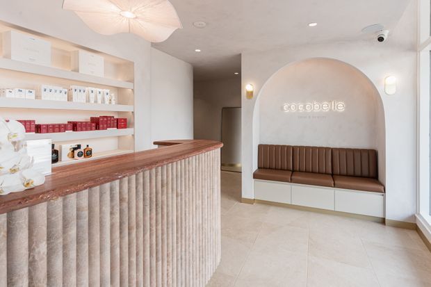 A refined beauty or retail reception interior with a curved fluted reception counter, pale tiled flooring, open product shelving, and a built-in brown upholstered bench set into a large arched wall niche. Soft cream and beige finishes, sculptural pendant lighting, and subtle signage create a calm, contemporary atmosphere.