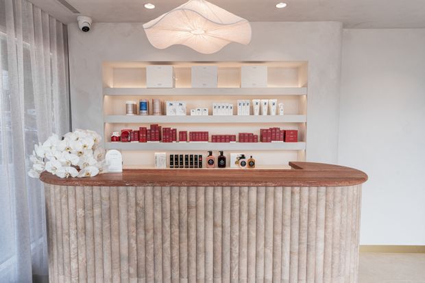 A refined retail or reception counter with a rounded, vertically fluted upholstered or textured front in warm beige tones and a dark timber benchtop. Illuminated open shelving behind displays neatly arranged skincare or beauty products, with soft floral pendant lighting, sheer curtains, recessed ceiling lights, and pale walls creating a calm, luxurious atmosphere.