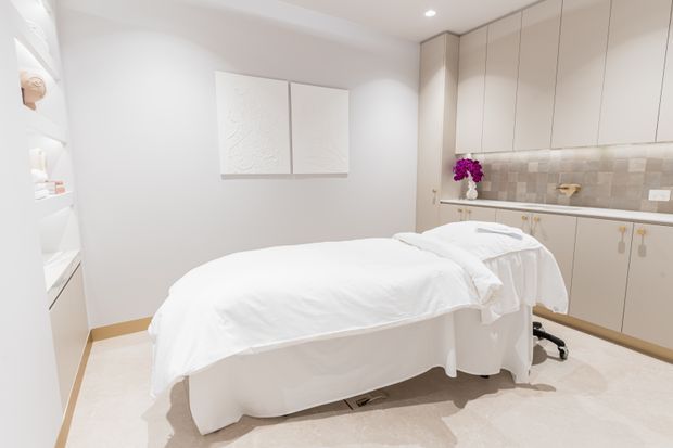 Bright, clean treatment room with two white treatment beds, pale flooring, curved white cabinetry, and a long wall of light beige built-in cabinets with a tiled splashback and countertop. Recessed lighting and minimal decor create a calm clinical-spa atmosphere.