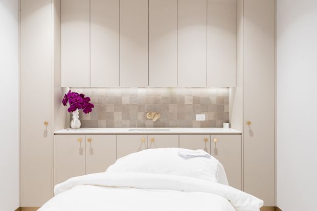 A compact, softly lit bedroom with a built-in bed set into a full-height wall of pale cabinetry. The bed has crisp white bedding, while a recessed backsplash niche behind the headboard features small square beige and taupe tiles, integrated lighting, a small outlet, and a decorative vase of purple flowers. Minimal hardware and concealed storage create a clean, tailored appearance.