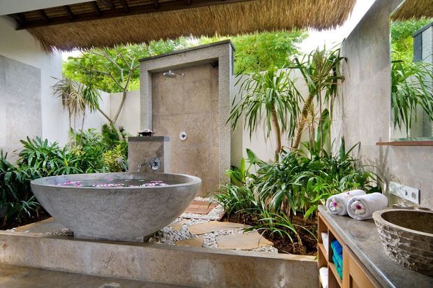 Open-air tropical bathroom with a freestanding stone soaking tub, exposed shower, stone basin, concrete-rendered walls, lush planting, pebble landscaping and a thatched roof canopy. The space has a resort-like, natural and tranquil character.