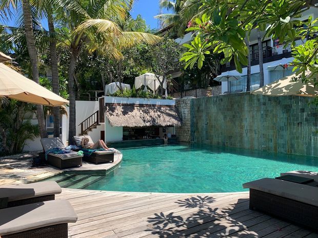 A tropical resort-style swimming pool courtyard with turquoise water, timber decking, lounge chairs, umbrellas, lush palms and frangipani planting. A textured tiled feature wall, shaded poolside structure and surrounding multi-level building create a relaxed outdoor setting.