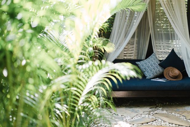 A tropical outdoor lounge nook viewed through lush foliage, featuring a dark built-in daybed or bench with patterned cushions and a straw hat. Sheer white curtains frame louvered windows behind the seating, while pale stone paving and white gravel create a relaxed resort-like setting.